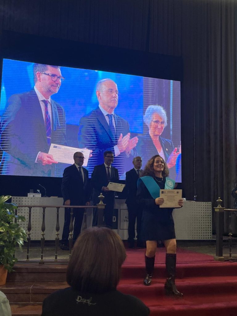 Una alumna titulada de la UNED Valencia sonríe con su diploma y beca (banda) tras recibir el reconocimiento durante el acto de apertura del curso 2025-2026.