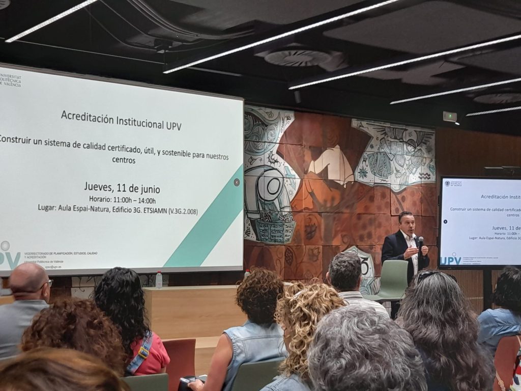 Un ponente interviene de pie junto a una gran pantalla que proyecta el título del evento 'Acreditación Institucional UPV: Construir un sistema de calidad certificado, útil y sostenible para nuestros centros'.