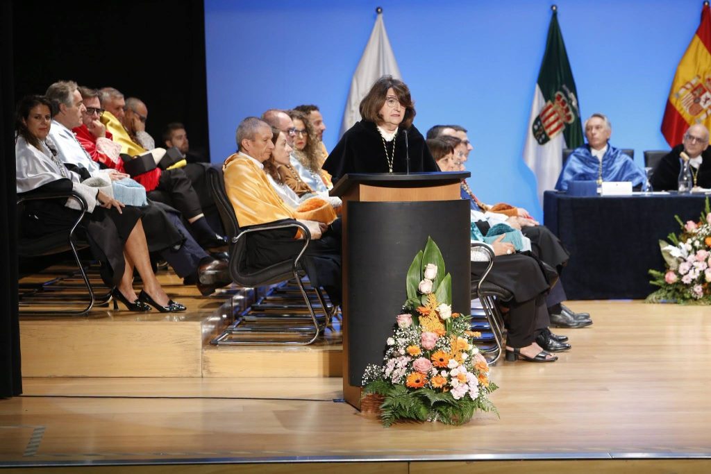 La rectora Amparo Navarro Faure pronuncia su discurso desde el atril con adorno floral durante el Solemne Acto de Apertura del Curso Académico 2026-2027 de la Universidad de Alicante, acompañada por autoridades e integrantes del claustro en el escenario.