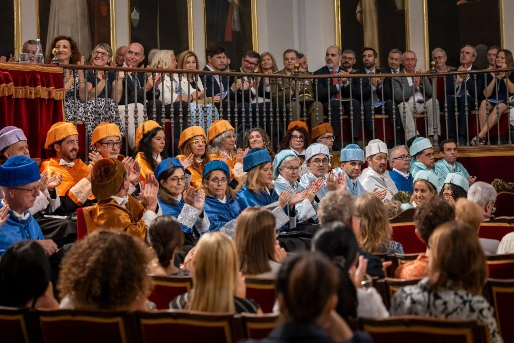 Asistentes y miembros del cuerpo docente e investigador con trajes académicos y birretes tradicionales presenciando la ceremonia de inicio de curso en el Paraninfo de la Universitat de València.